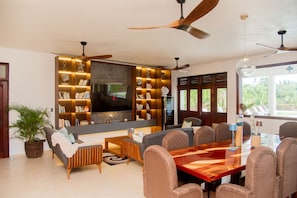 A modern living room with a large flat-screen TV, built-in bookshelves, a dining area with a wooden table, and a ceiling fan.