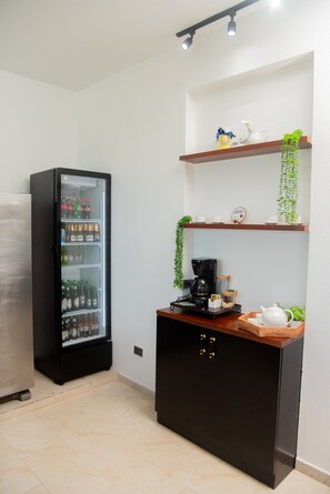 A kitchenette with a refrigerator stocked with beverages, a coffee maker, and a tea set on a wooden counter.