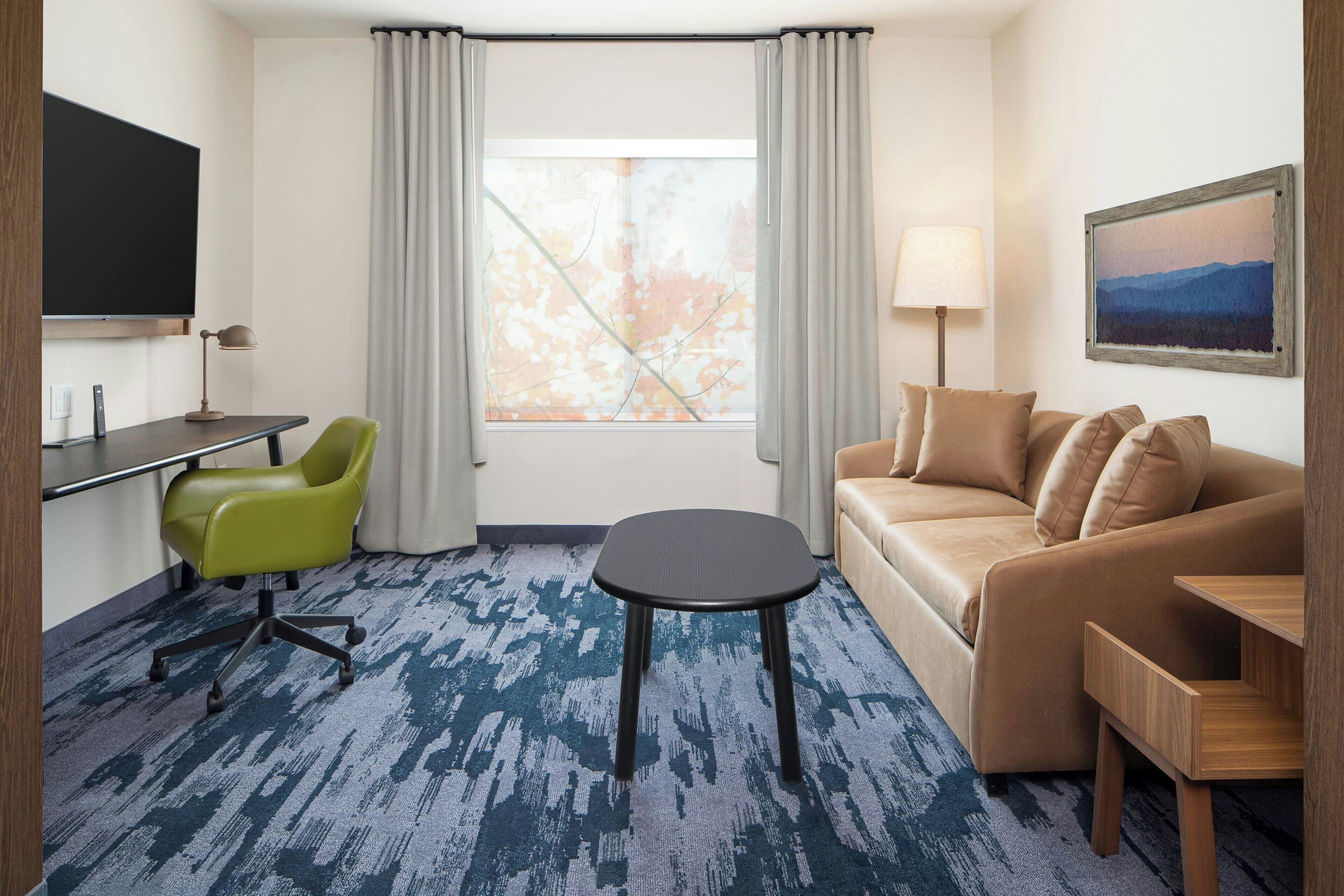 A hotel room with a sofa, a desk with a chair, a TV, a lamp, and a painting on the wall.