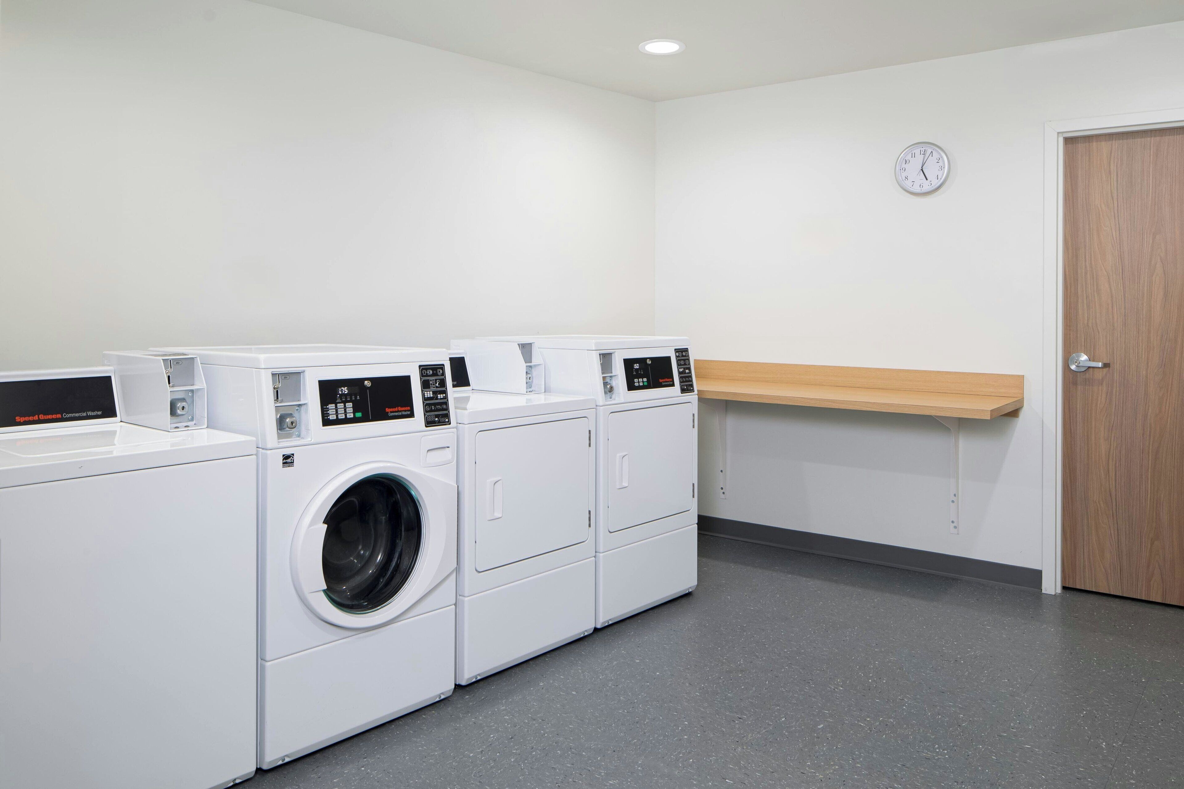 A laundry room with multiple washing machines and dryers, a clock on the wall, and a wooden shelf.