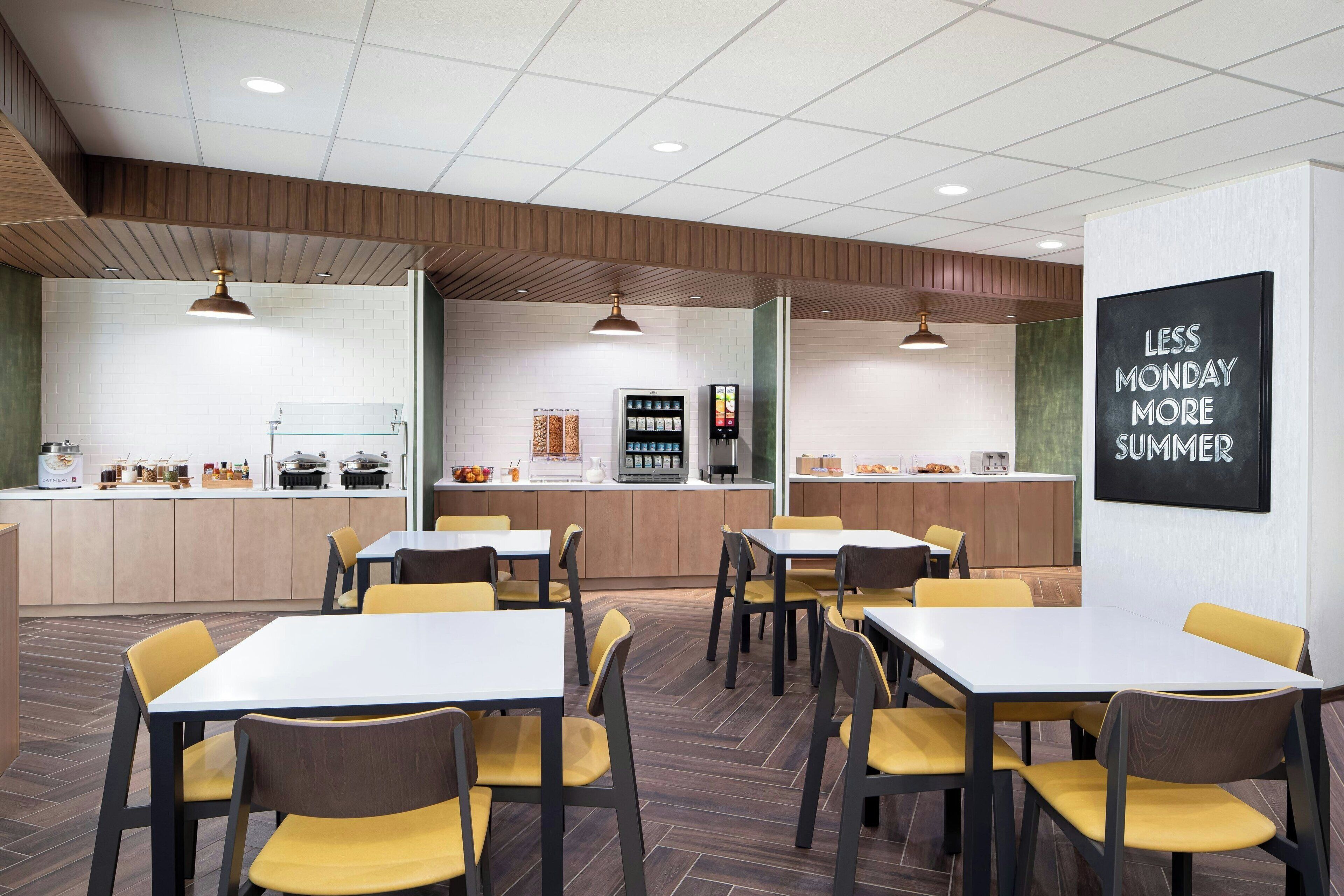 Una cafetería moderna con mesas y sillas, una zona de cocina y un cartel motivacional en la pared.