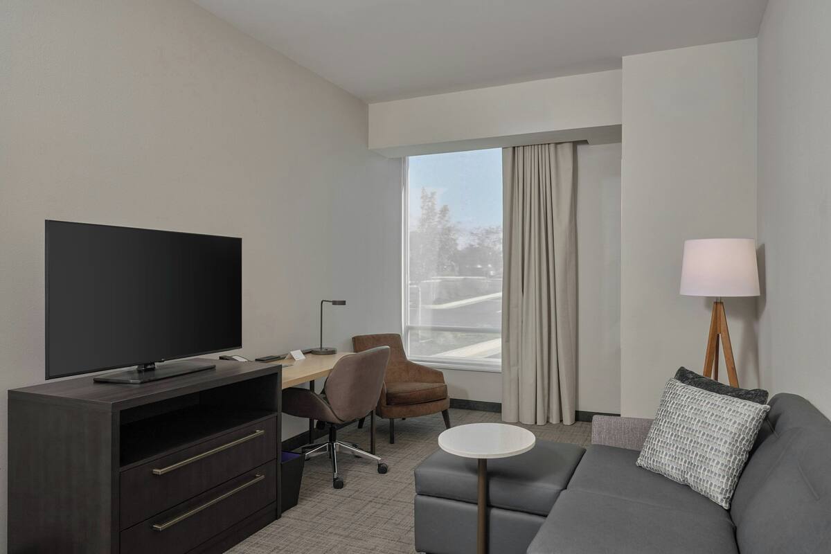 Una habitación de hotel moderna con televisión de pantalla plana, un escritorio con silla, un sofá, una mesita redonda y una lámpara.