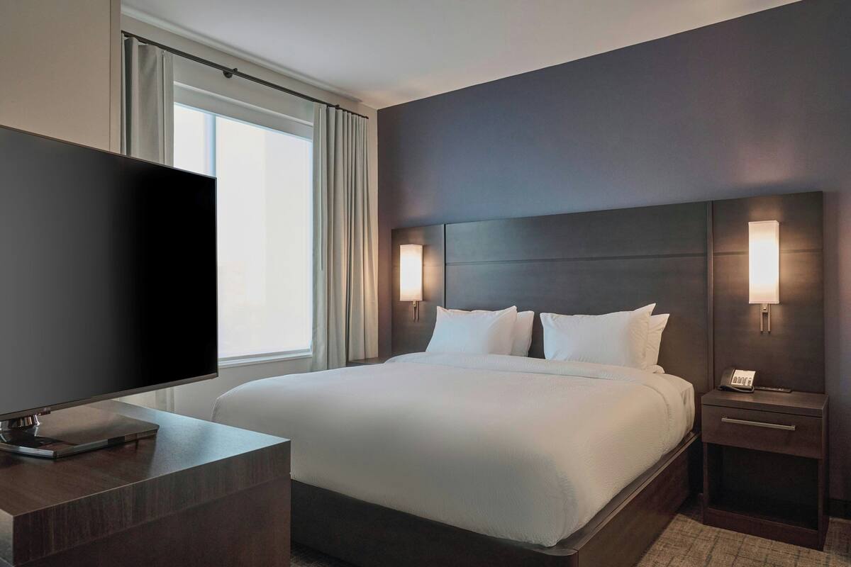 Una habitación de hotel moderna con una cama grande, un televisor de pantalla plana, una mesita de noche y un atril con un teléfono.