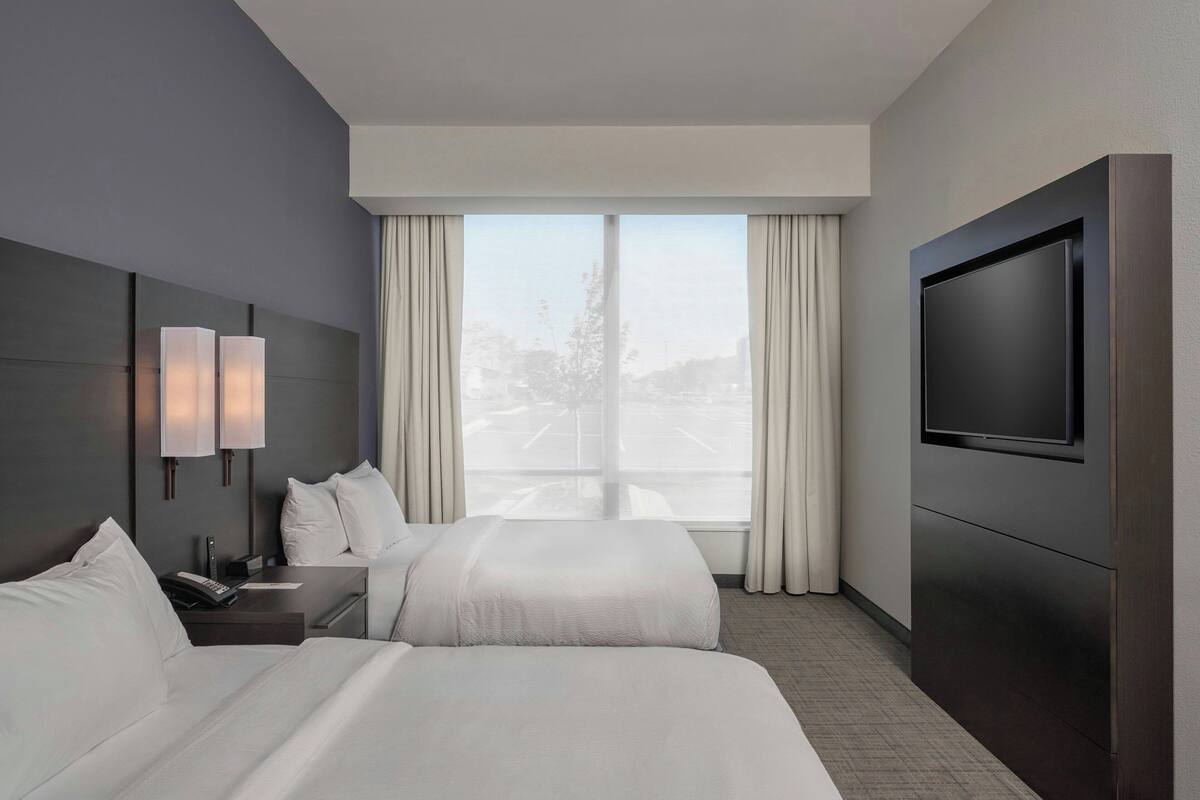 Habitación de hotel con dos camas, un televisor de pantalla plana, una mesita de noche y una ventana con cortinas.
