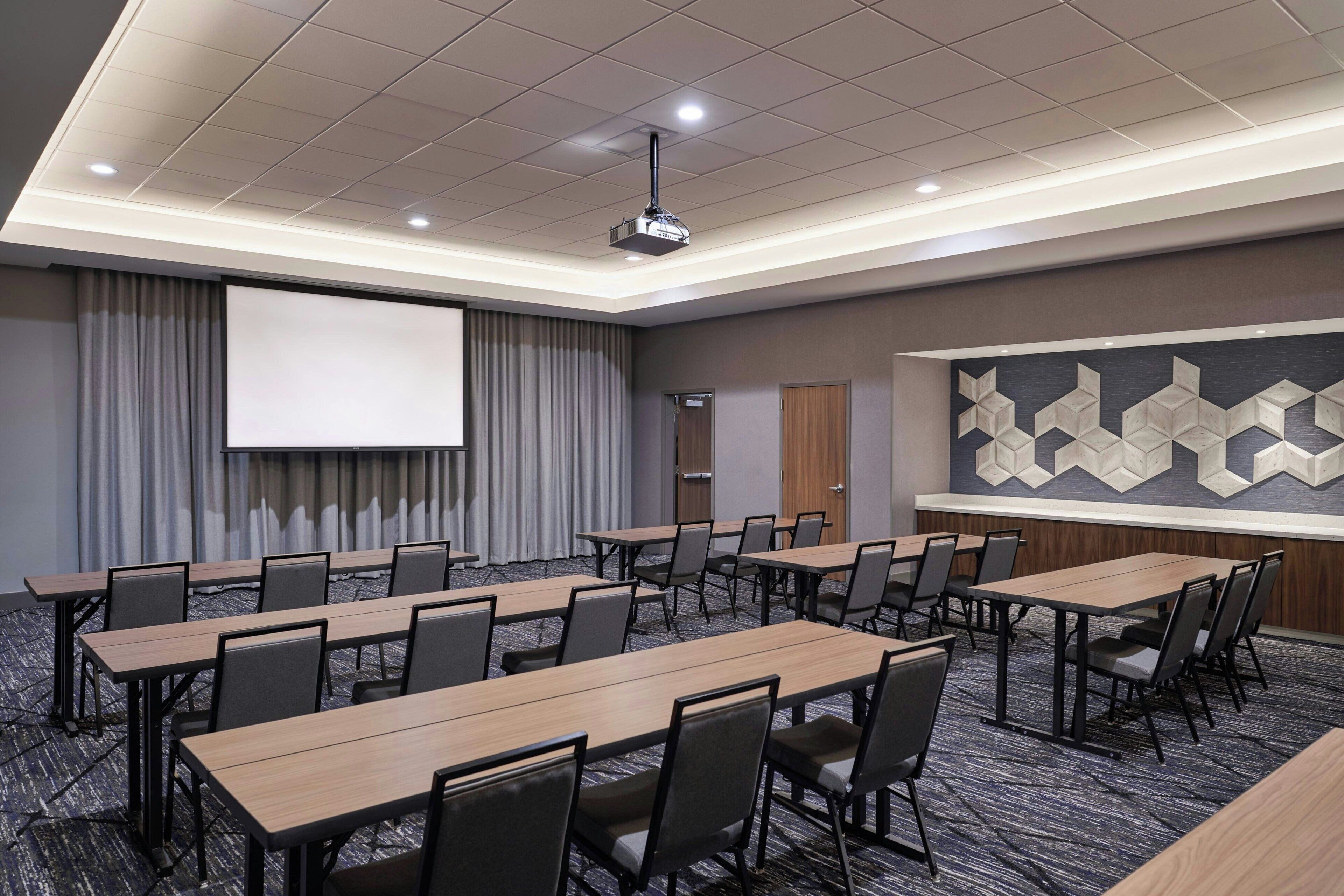 A conference room with multiple tables and chairs, a projector screen, and a large geometric wall decoration.