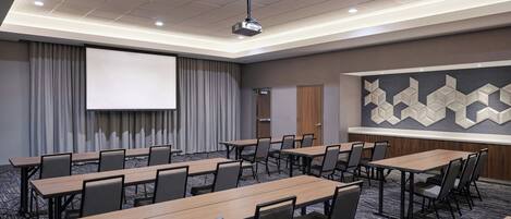 A conference room with multiple tables and chairs, a projector screen, and a large geometric wall decoration.