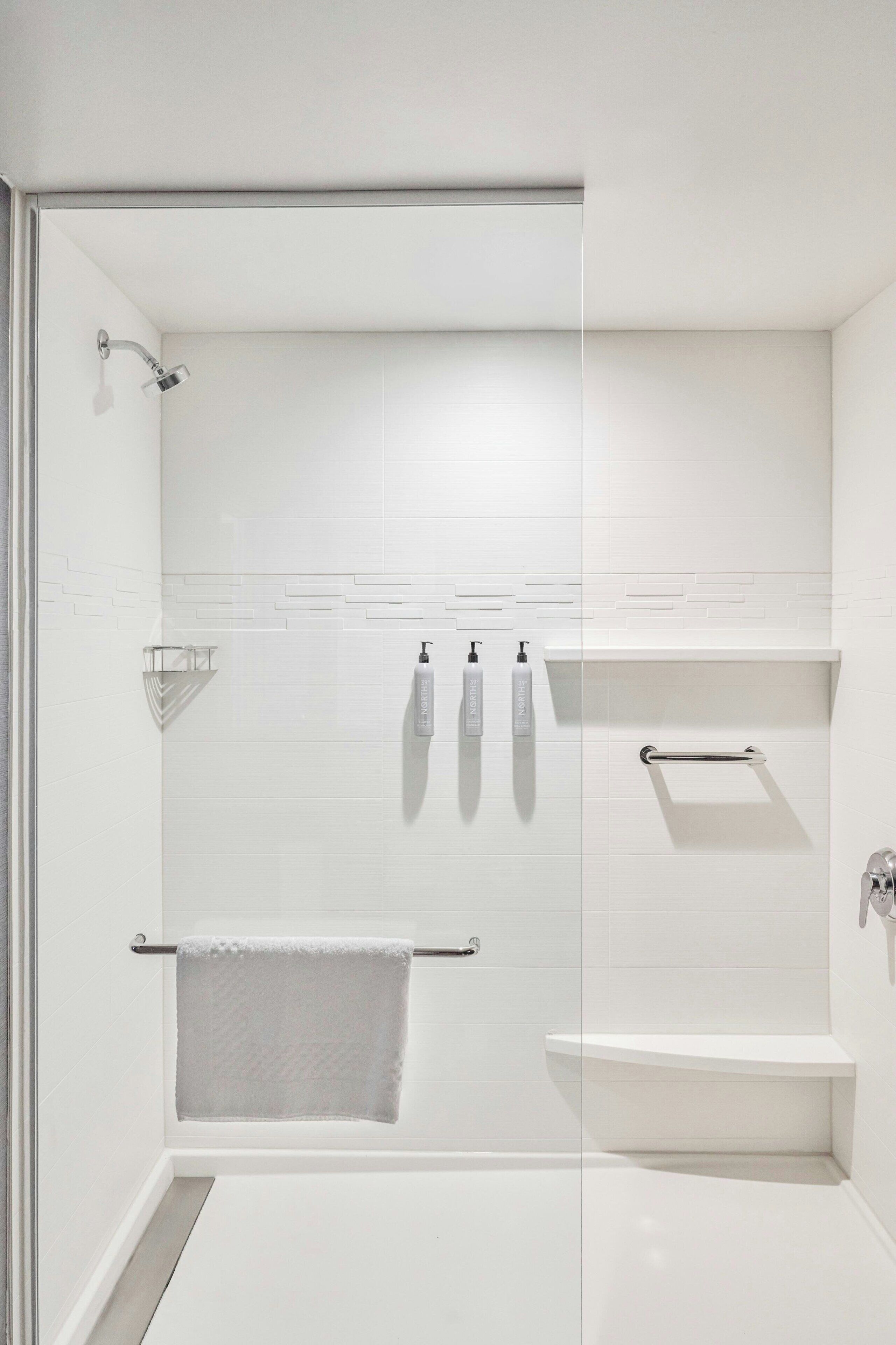 Un baño moderno con cabina de ducha de vidrio, paredes cubiertas de azulejos blancos y un estante empotrado con artículos de aseo.