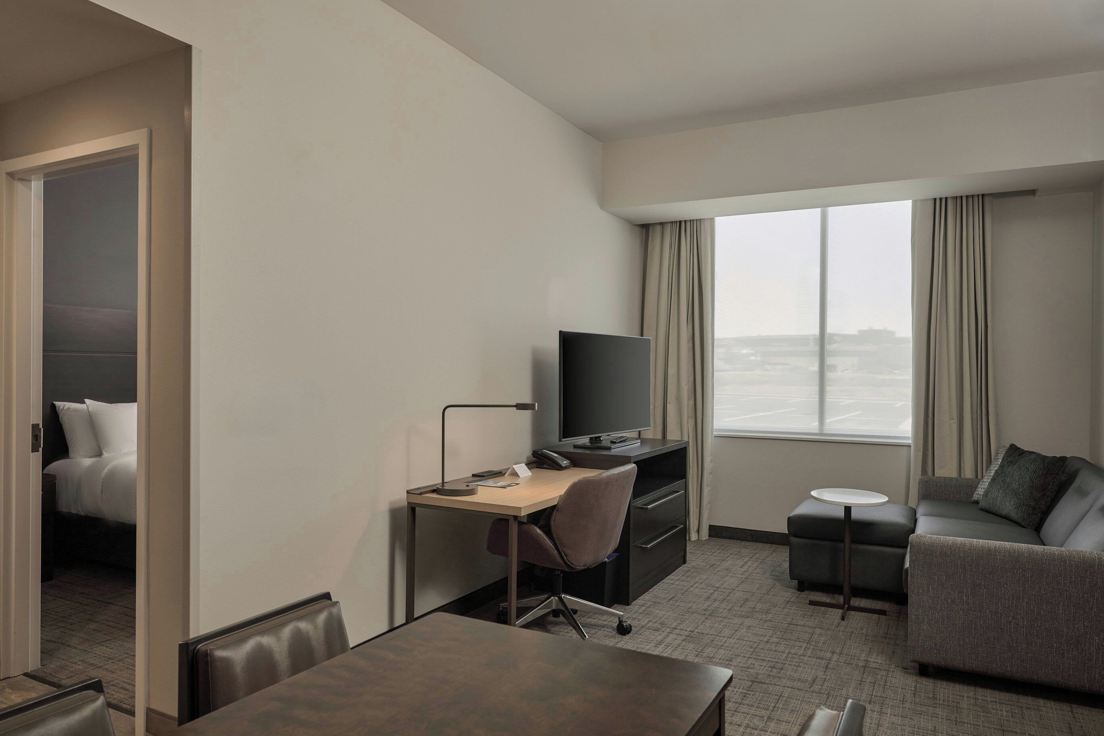 A hotel room with a bed, a desk with a computer, a television, a sofa, and a window with a view of the city.