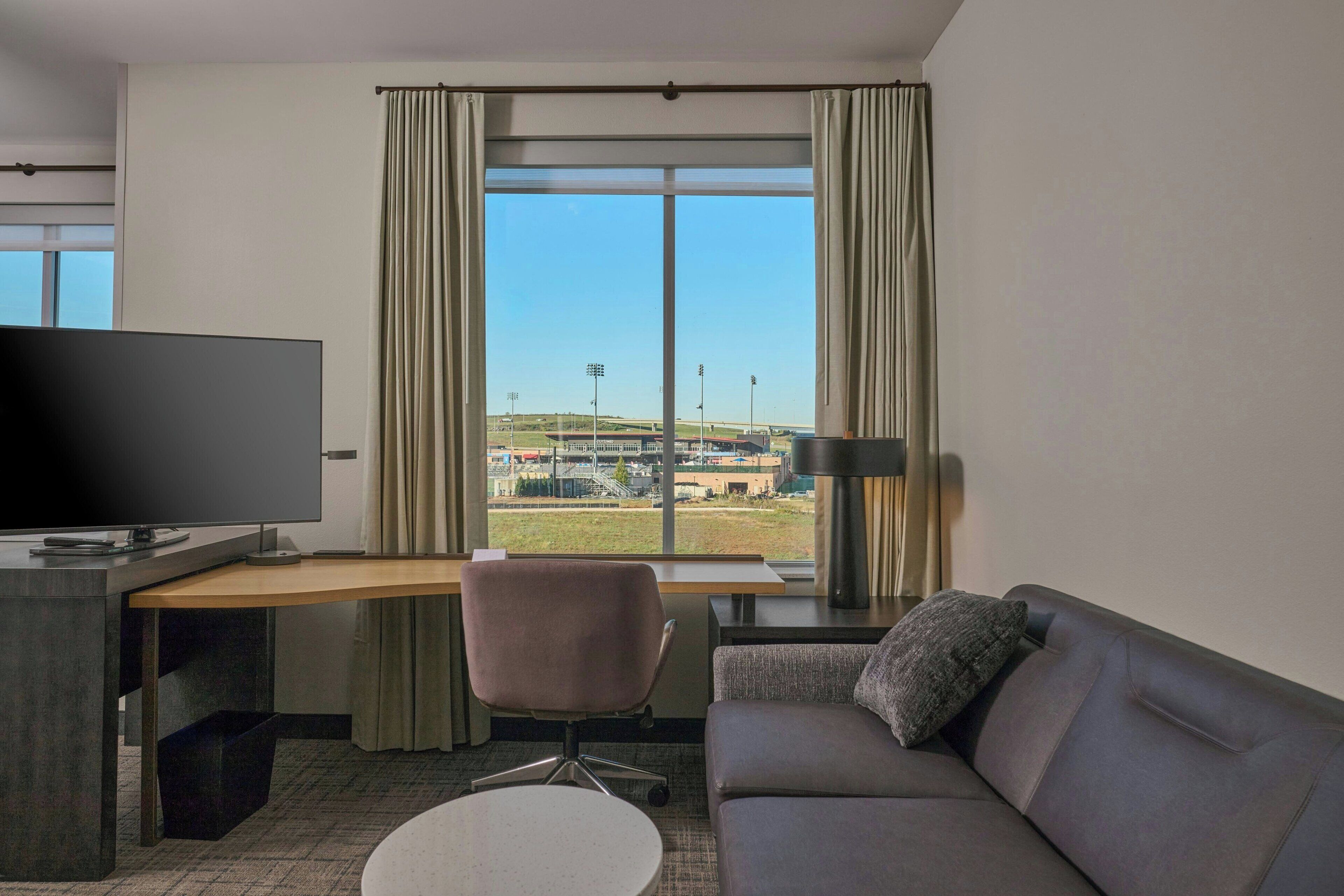 A hotel room with a large window, a desk, a television, a sofa, and a lamp.