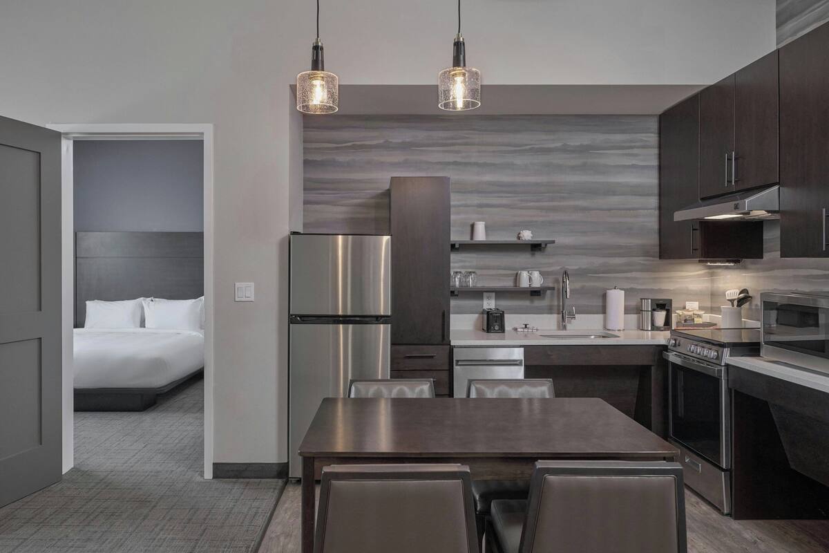 Una cocina moderna con electrodomésticos de acero inoxidable, una zona de comedor con mesa y sillas, y un dormitorio visible a través de un umbral abierto.