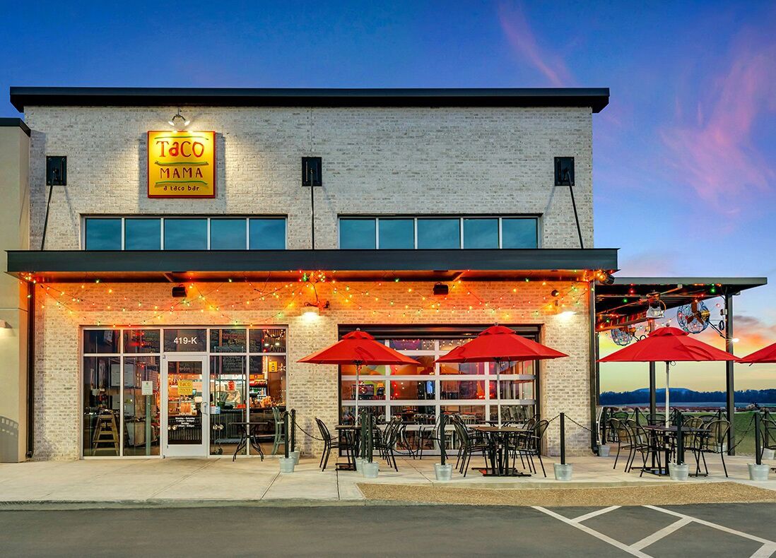 A restaurant with a brick facade, outdoor seating with red umbrellas, and a sign reading "Taco Mama".