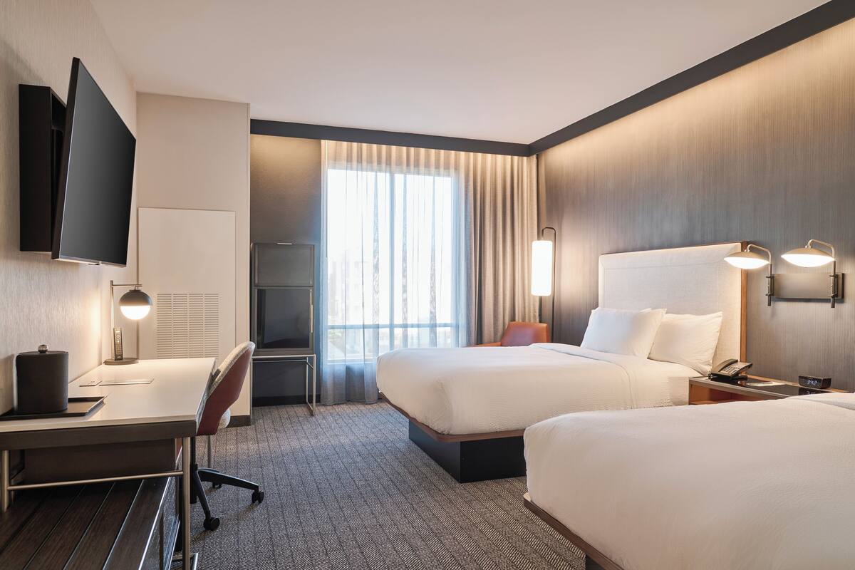 A hotel room with a desk, two beds, a TV, and a large window with curtains.