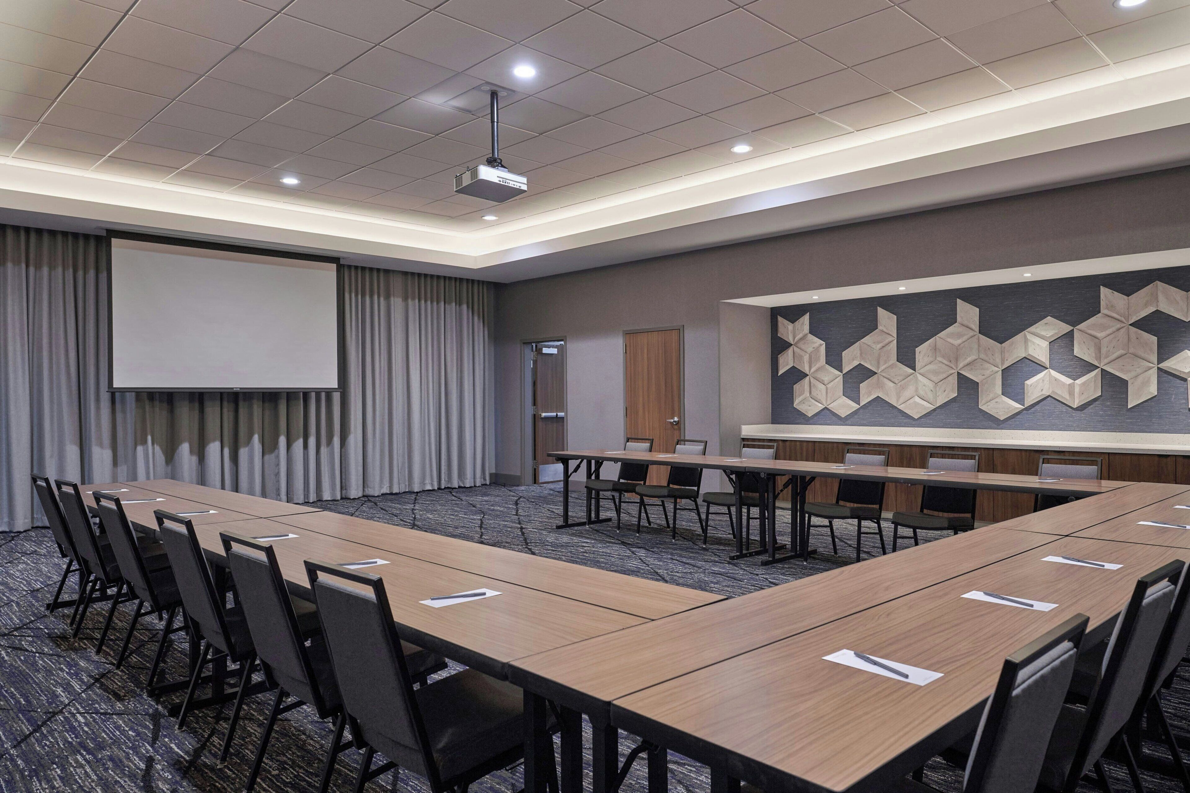 A conference room with a long table, chairs, a projector screen, and a patterned wall.
