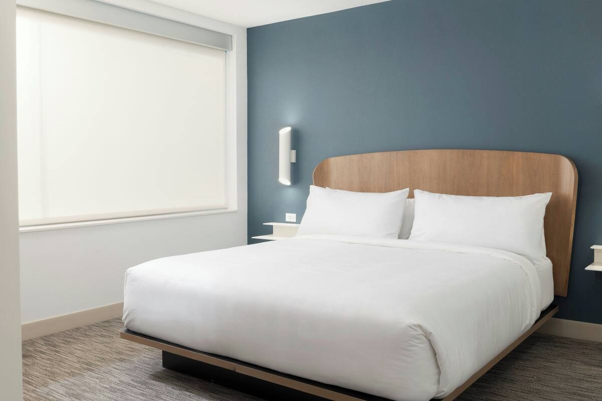 Ein modernes Hotelzimmer mit einem großen Bett, einem Nachttisch, einer Wandleuchte und einem Fenster mit Jalousien.