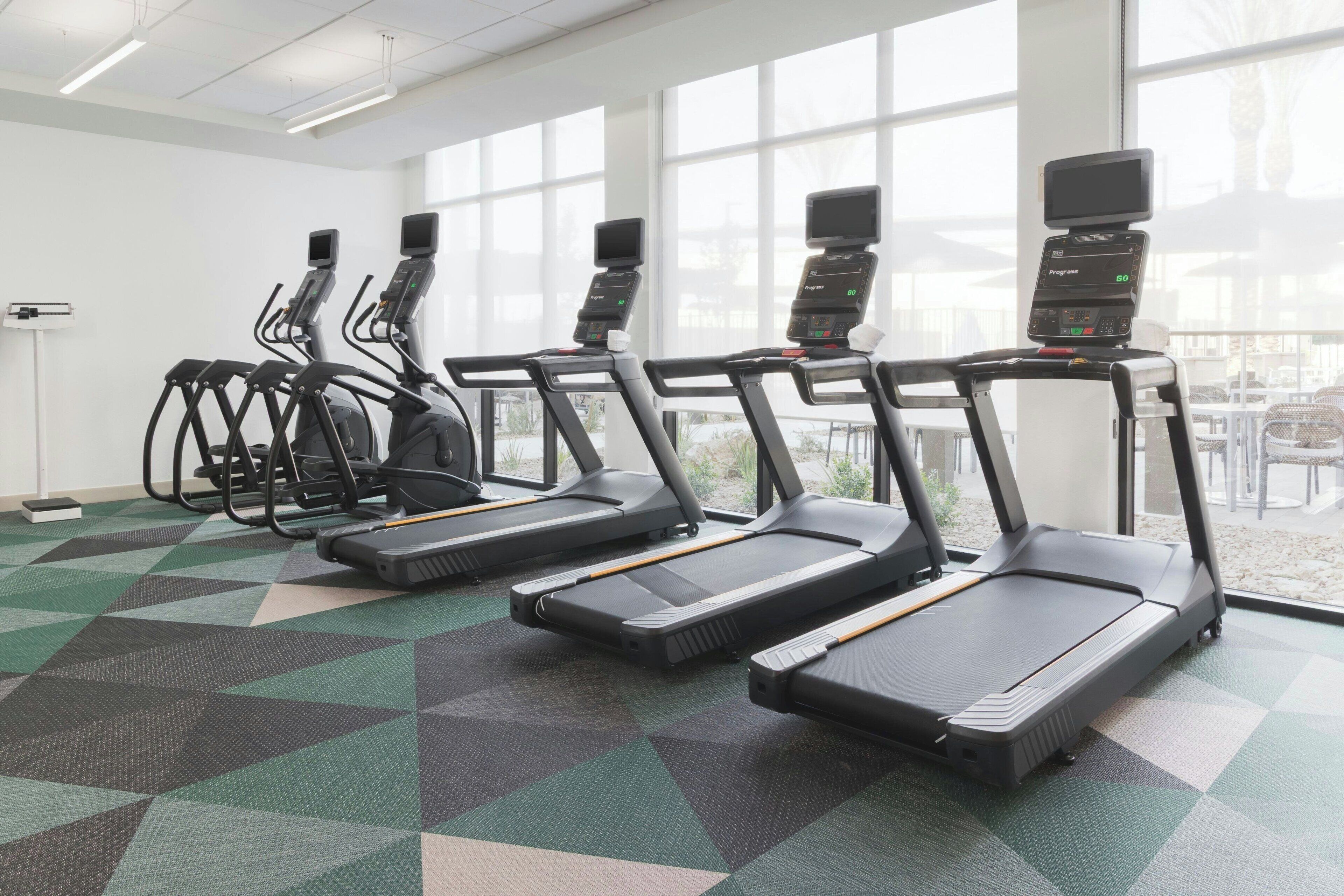 Une salle de sport avec des tapis de course alignés en rangées, de grandes fenêtres et un sol à motifs.