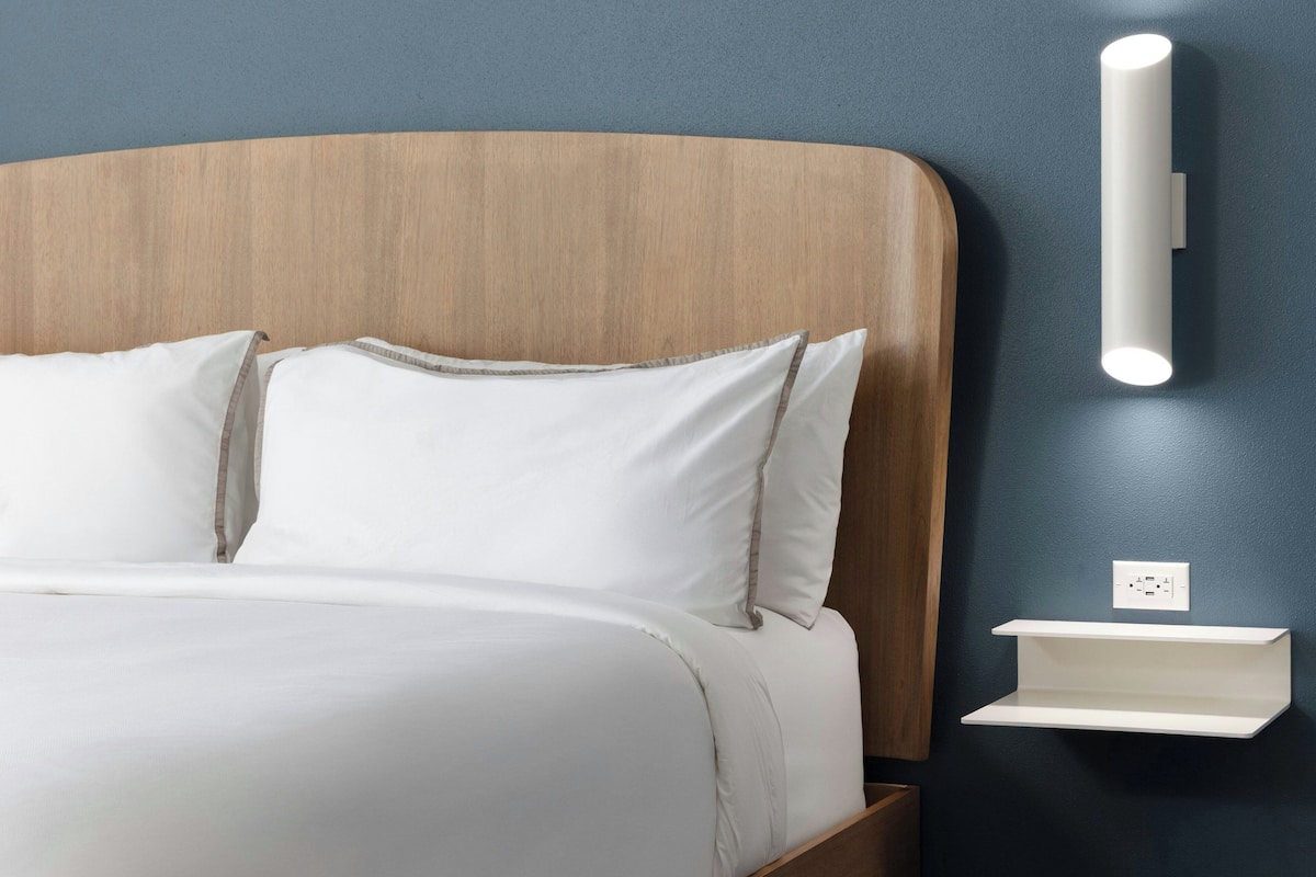 Ein Bett mit weißer Bettwäsche, ein Holz-Kopfstück und ein Nachttisch in Weiß mit einer Wandleuchte.