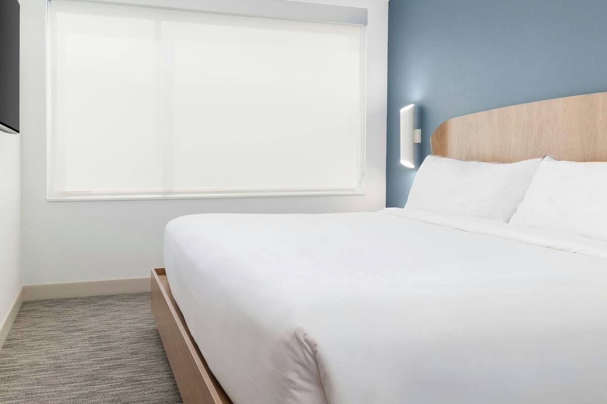 Ein ordentlich bezogenes Bett mit weißen Leinen in einem Zimmer mit blauer Akzentwand und einem Fenster mit Jalousien.