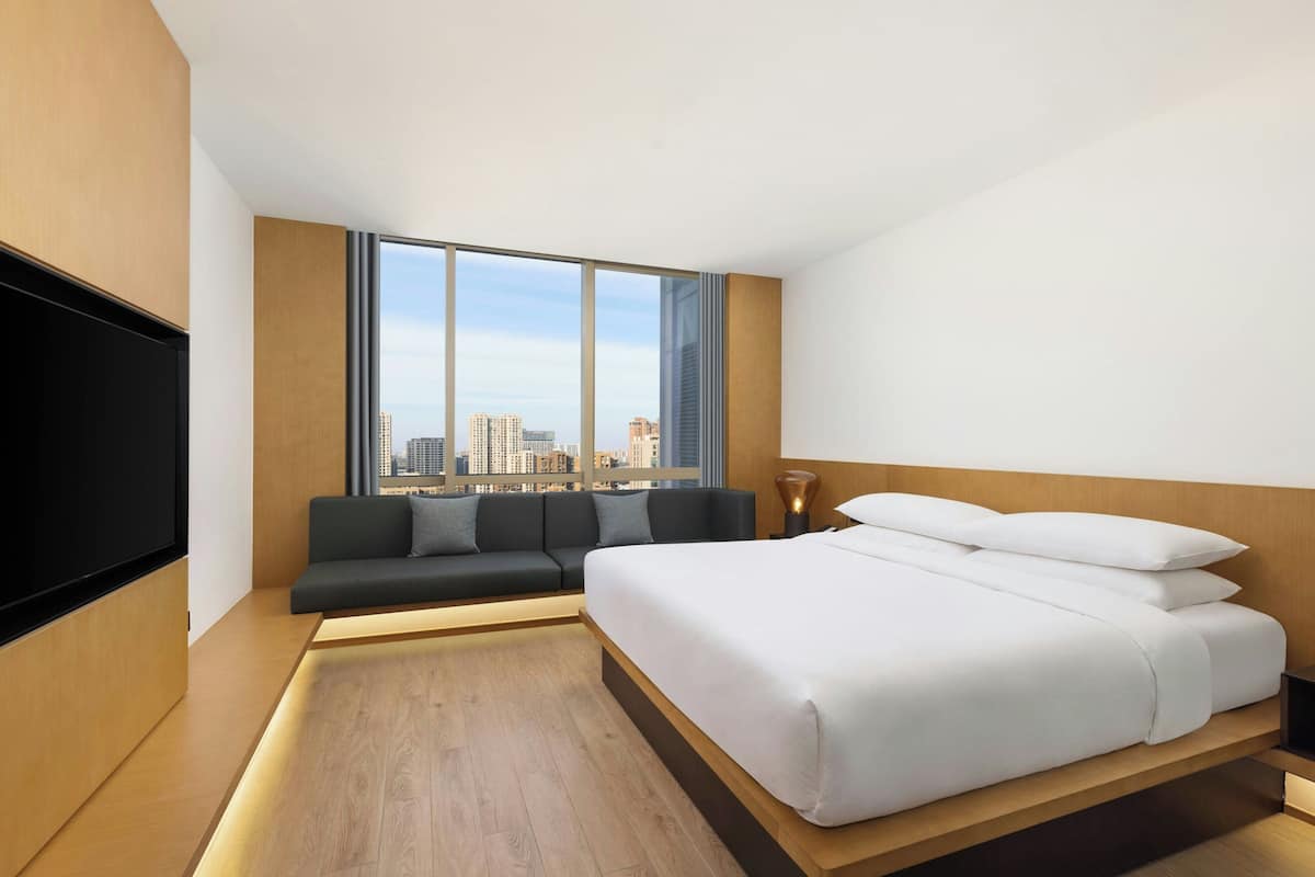 A modern hotel room with a large bed, a sofa, a TV, and a city view.