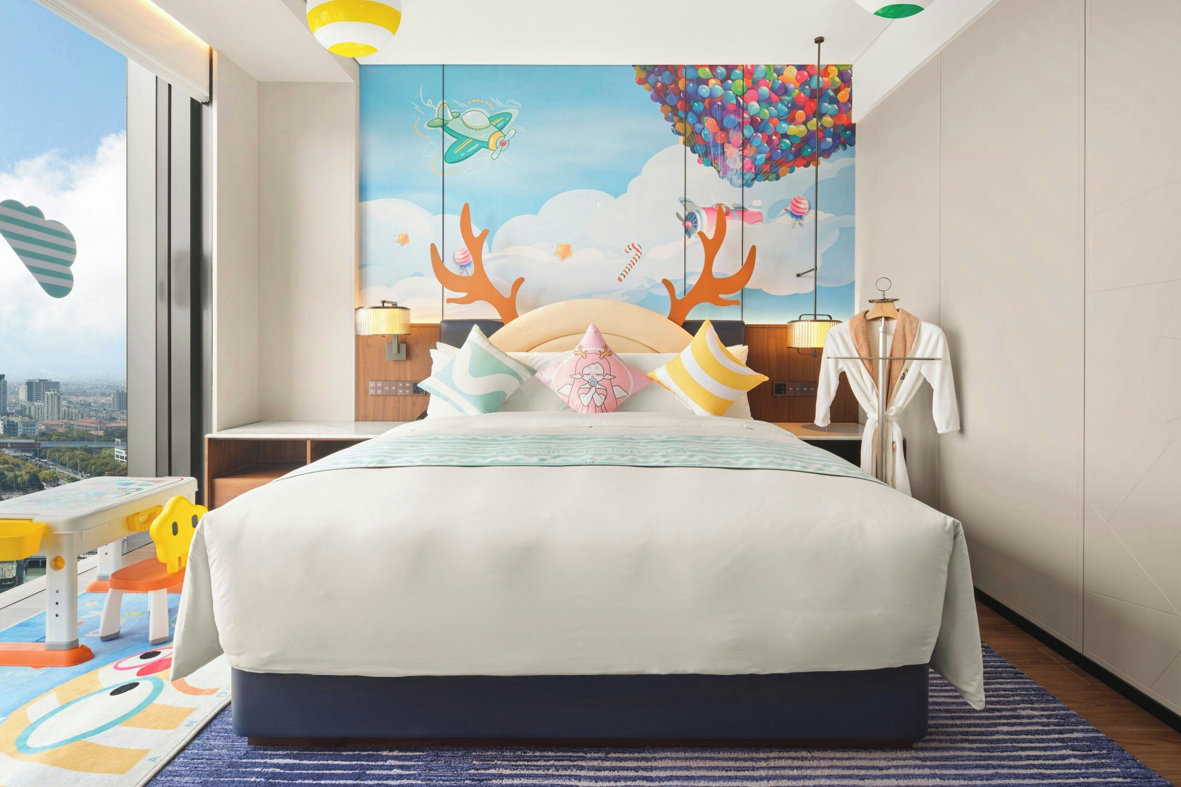 A hotel room with a large bed, a city view, a mural with a flying airplane and balloons, and a wooden nightstand with a lamp.