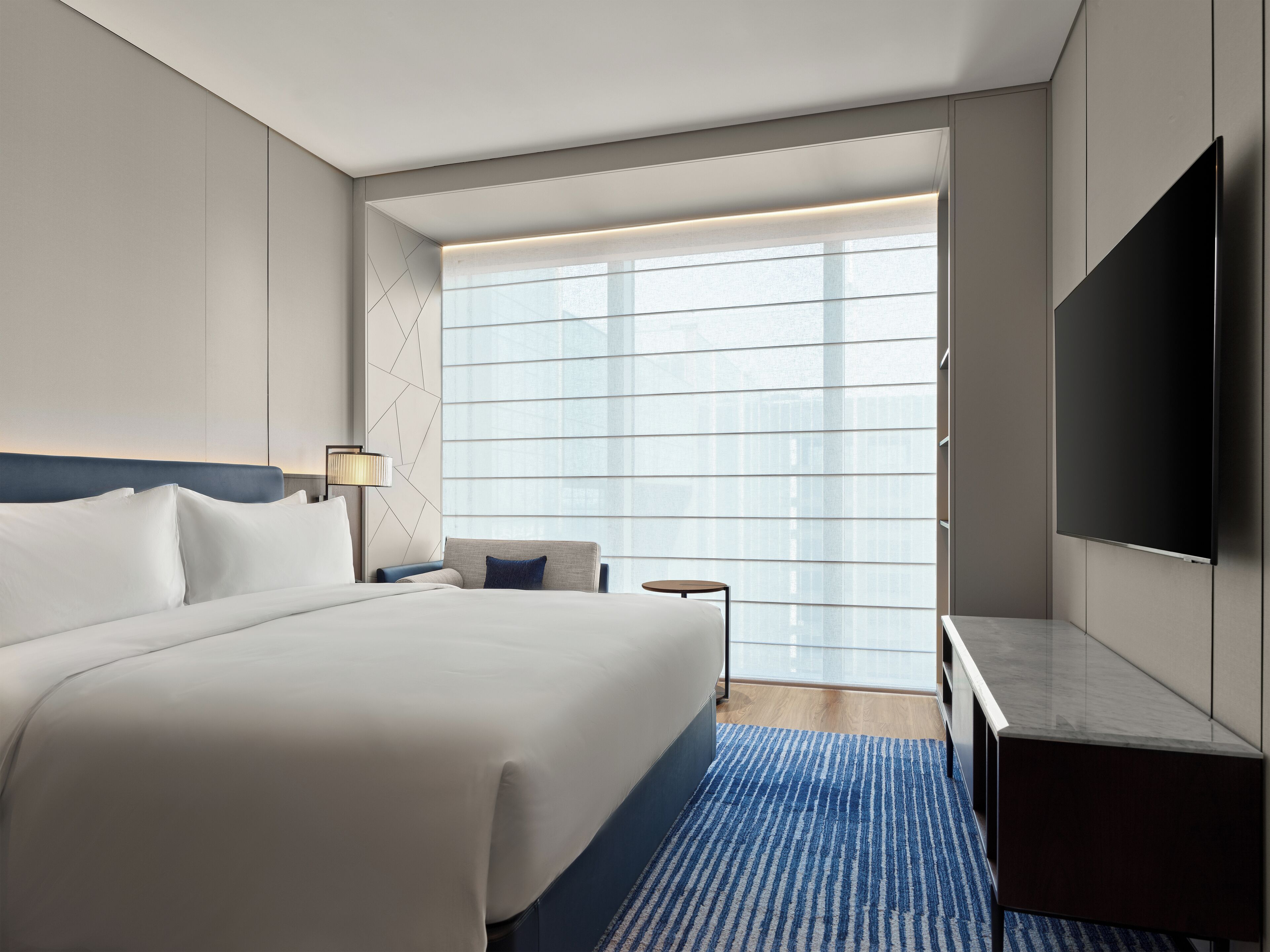 A modern hotel room with a large bed, a desk, a TV, and a window with blinds.