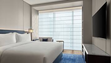 A modern hotel room with a large bed, a desk, a TV, and a window with blinds.