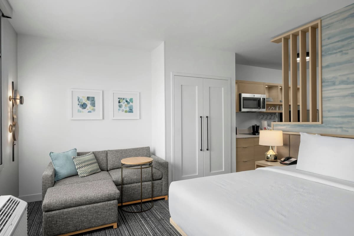Une chambre d’hôtel moderne avec un canapé gris, une table ronde, un lit, un four à micro-ondes et un petit coin cuisine.