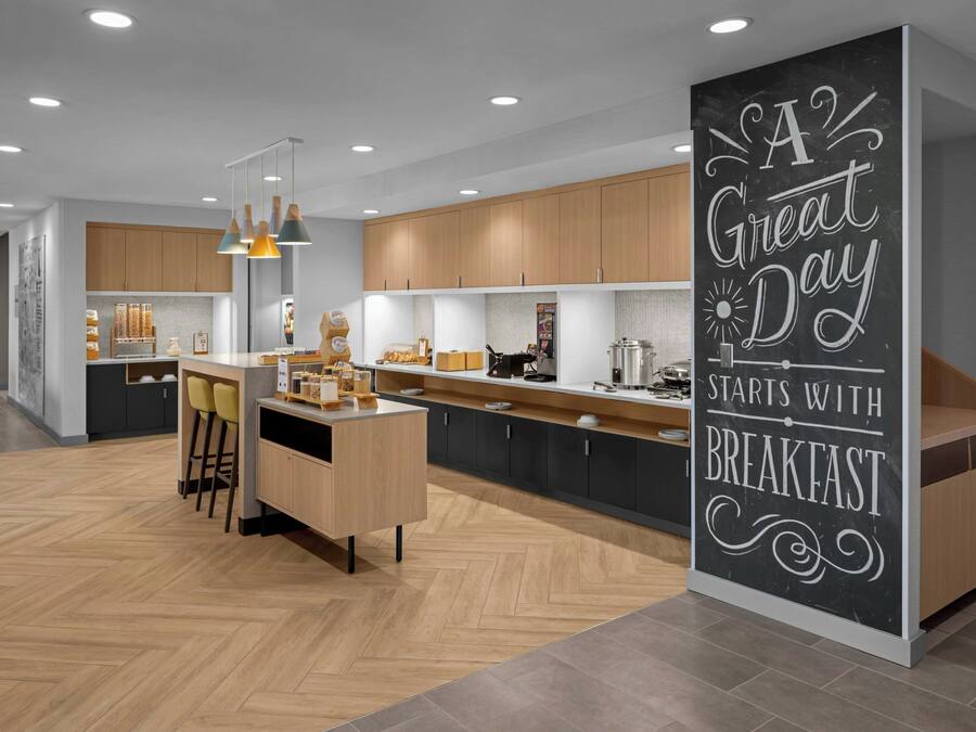 A modern kitchen with wooden cabinets, a central island with bar stools, pendant lighting, and a chalkboard sign saying "A Great Day Starts with Breakfast".