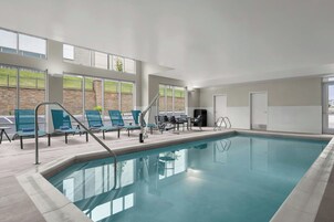 An indoor swimming pool with clear blue water, surrounded by lounge chairs and a sun terrace with a view of a grassy area.