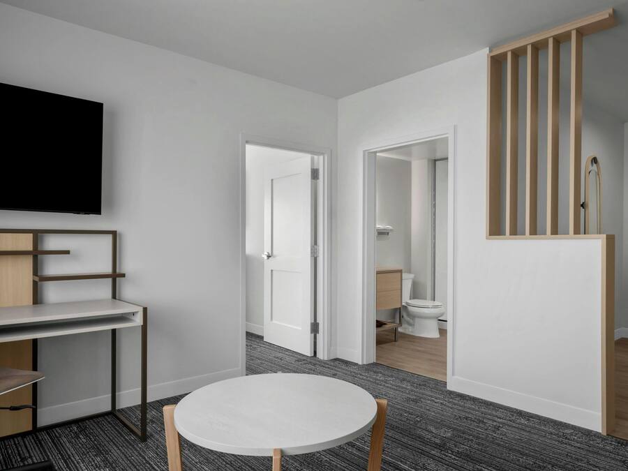 Una habitación de hotel moderna con una televisión de pantalla plana, un escritorio con silla, una mesa redonda y una escalera con barandillas de madera.