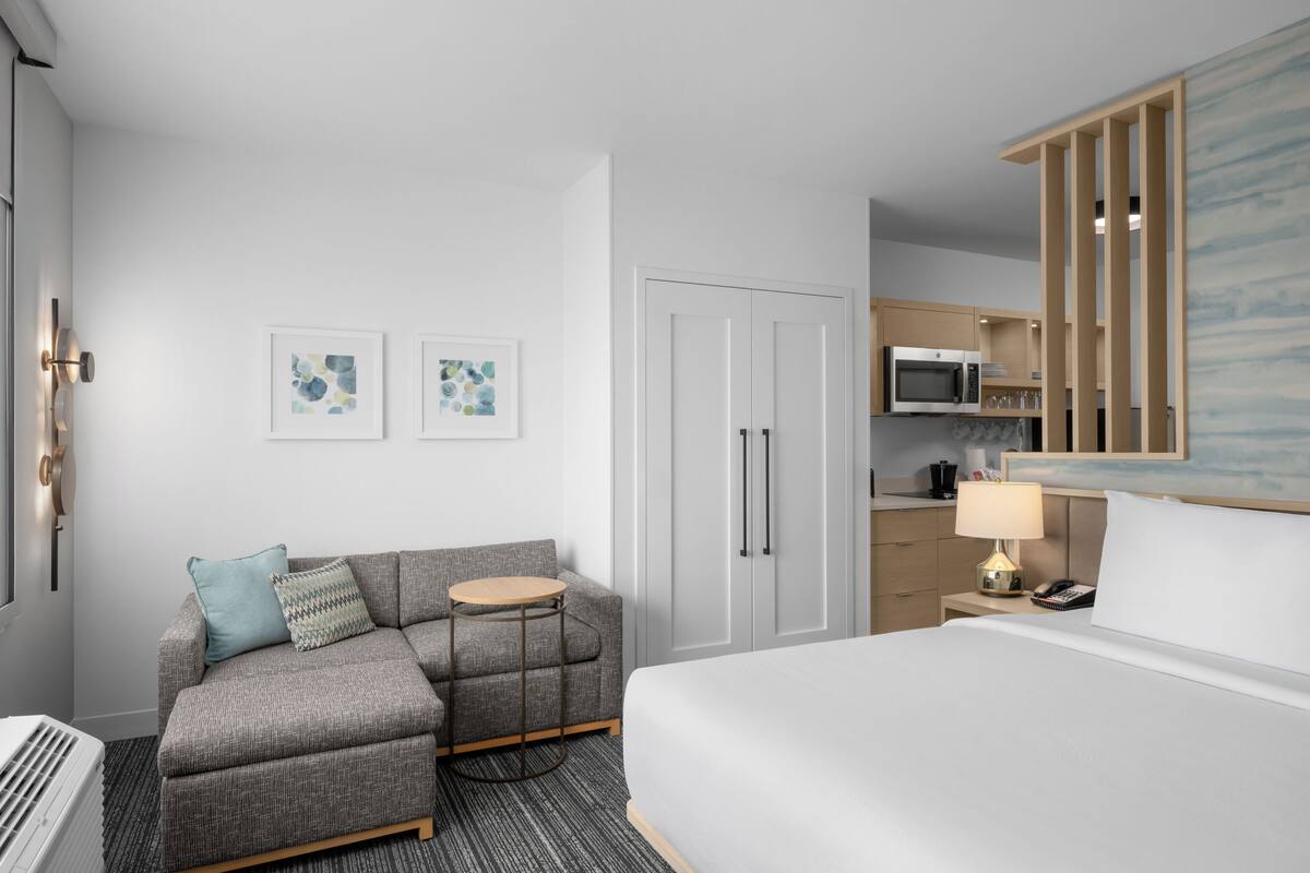 Une chambre d’hôtel moderne avec un canapé gris, une petite table ronde, un lit, un coin cuisine et une porte blanche.