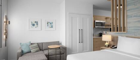 A modern hotel room with a grey sofa, a small round table, a bed, a kitchenette, and a white door.