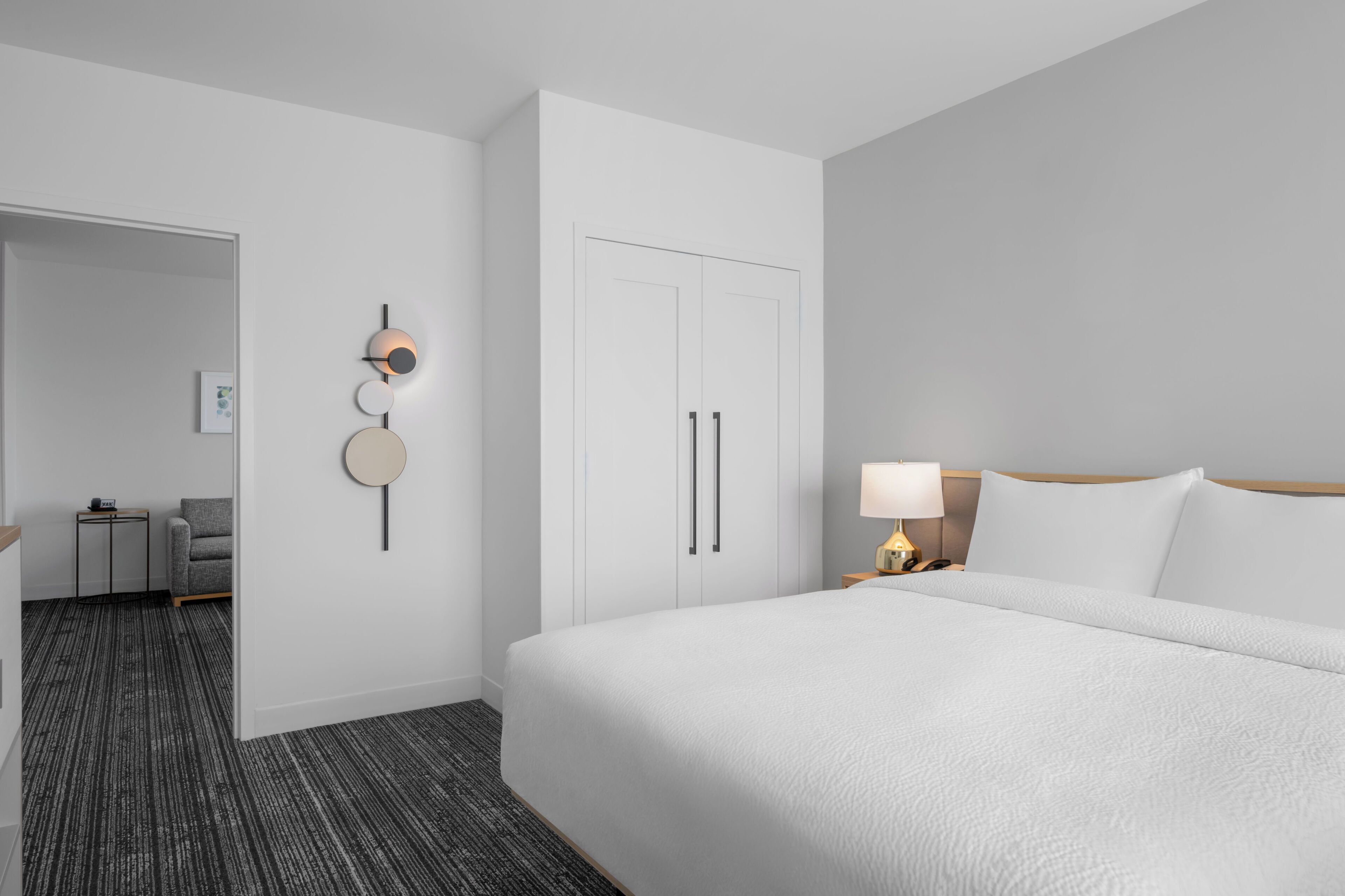 A hotel room with a large bed, a bedside table with a lamp, a wardrobe, and a small seating area with a chair and a table.