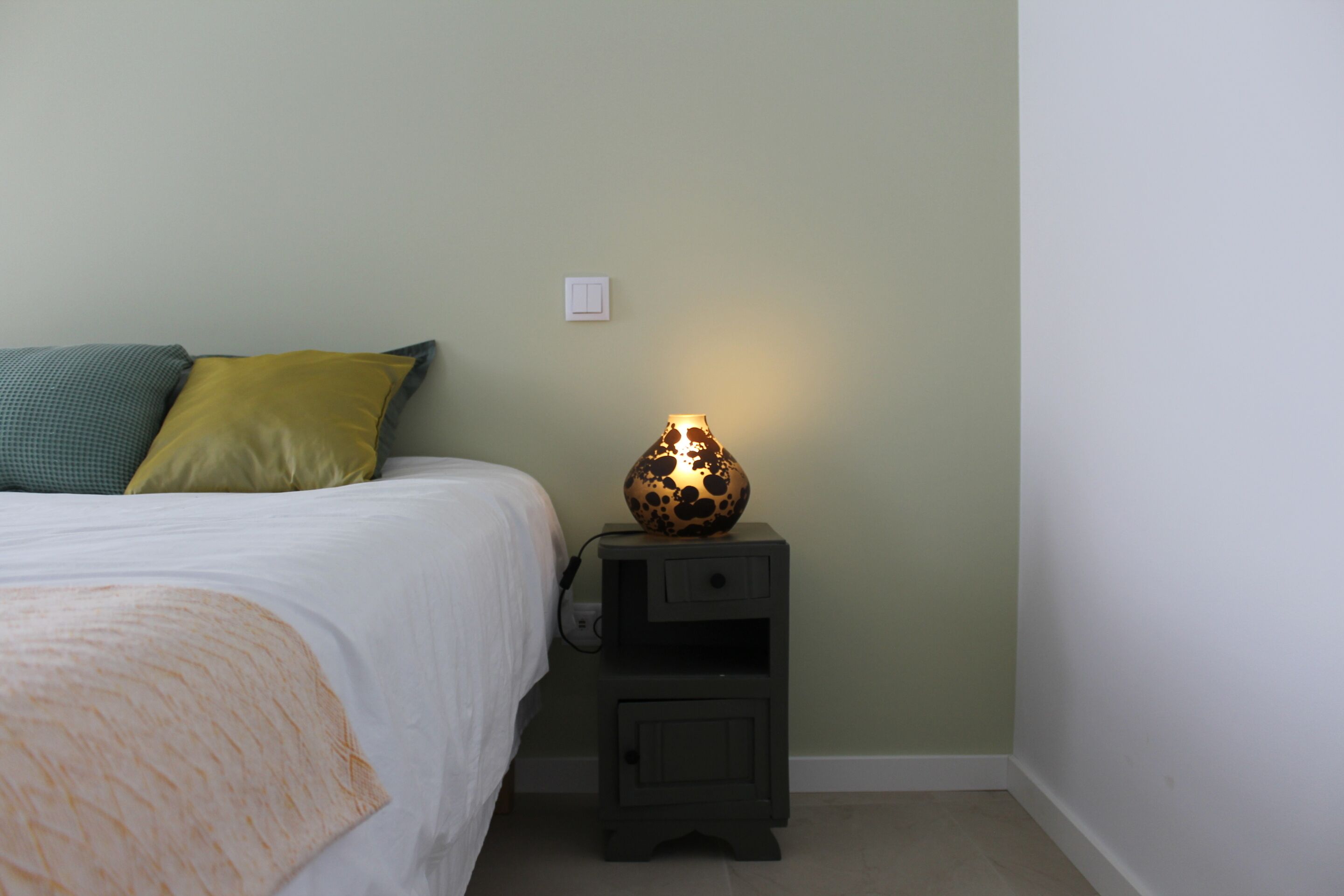 A bedroom with a bed, a nightstand with a lamp, and a wall switch.