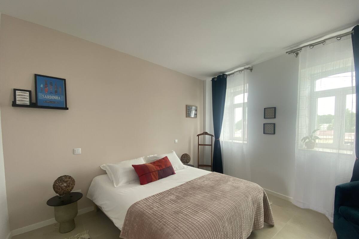 Un dormitorio con cama, mesita de noche, una silla, ventana con cortinas y adornos en la pared.