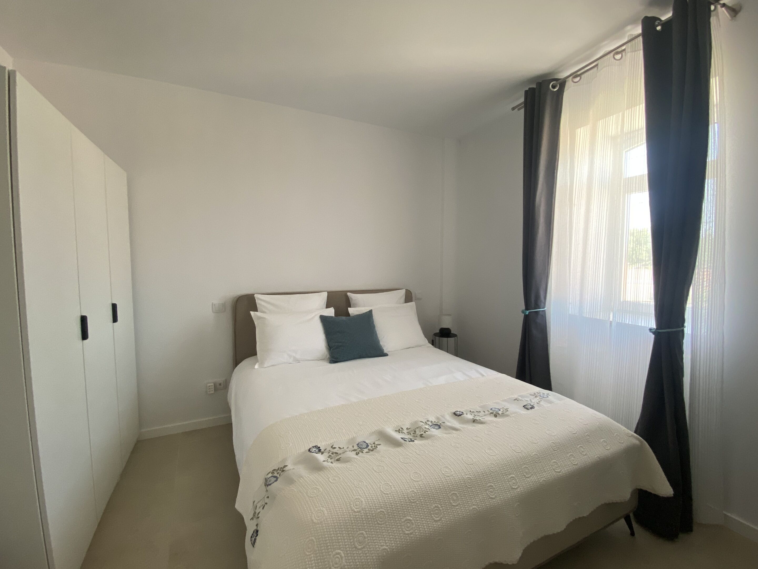 A bedroom with a bed, a nightstand, a wardrobe, and a window with curtains.