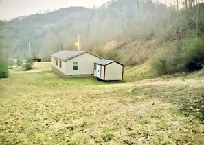 Une maison de plain-pied avec un petit garage attenant, entourée d’une cour verdoyante et d’une colline boisée.