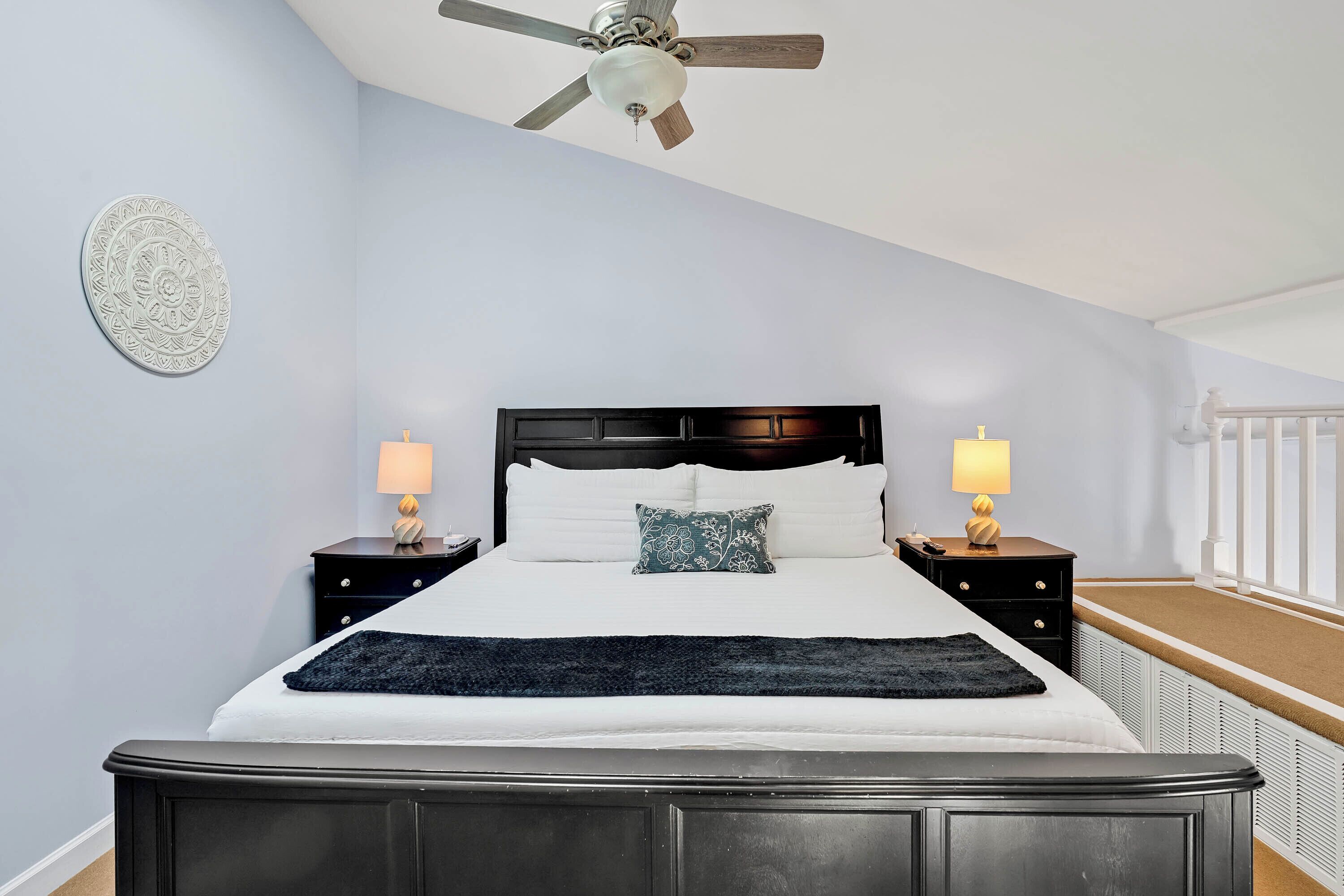 A bedroom with a bed, two bedside tables, a ceiling fan, a decorative wall plate, and a window with a white railing.