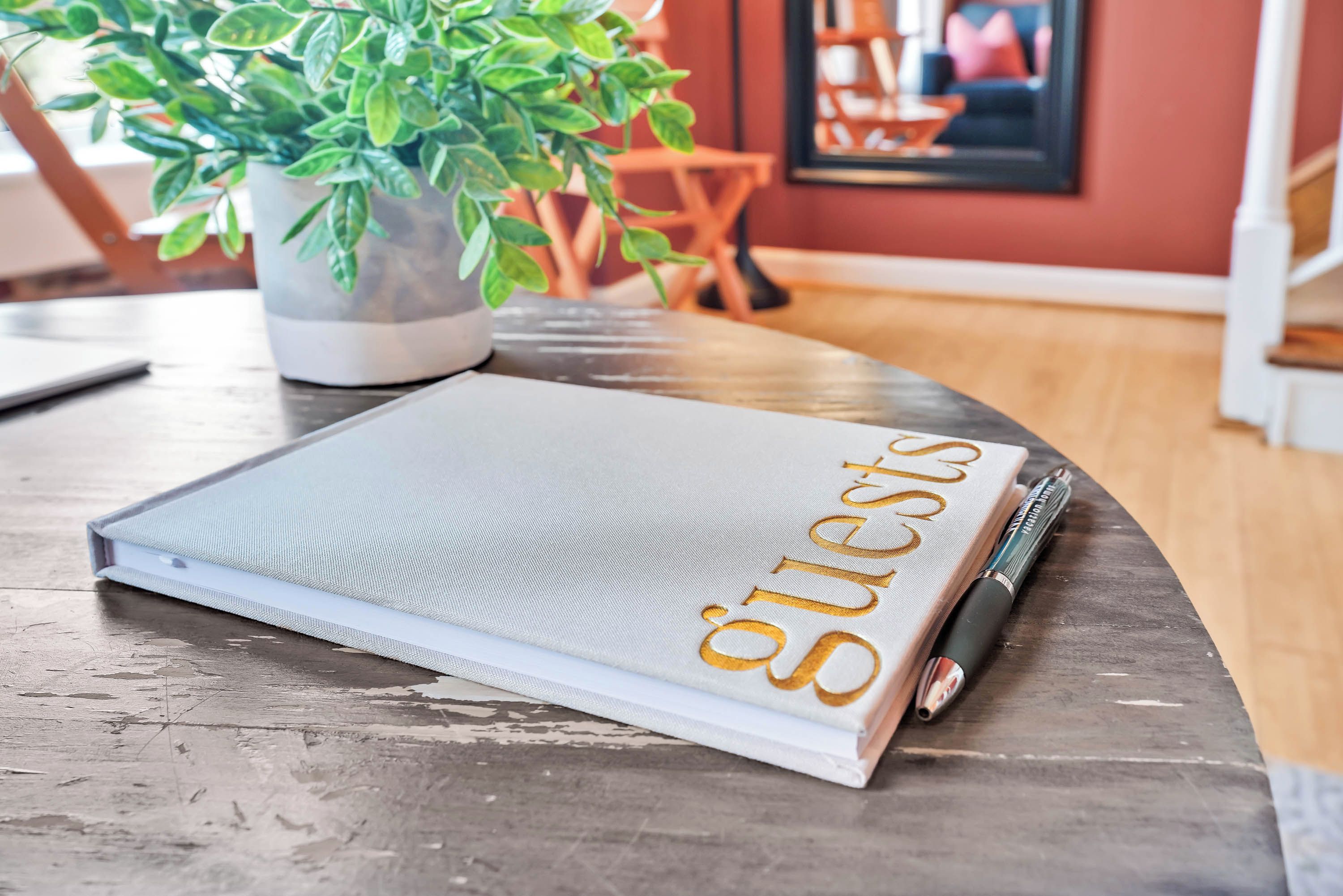 Een boek met de titel 'Guests' op een houten tafel, met een potplant en een lijst met foto in de achtergrond.