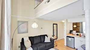 Ein modernes Wohnzimmer mit einem dunkelgrauen Sofa, einem kleinen Beistelltisch und einer Küchenzeile inklusive Geschirrspüler und Spiegel.