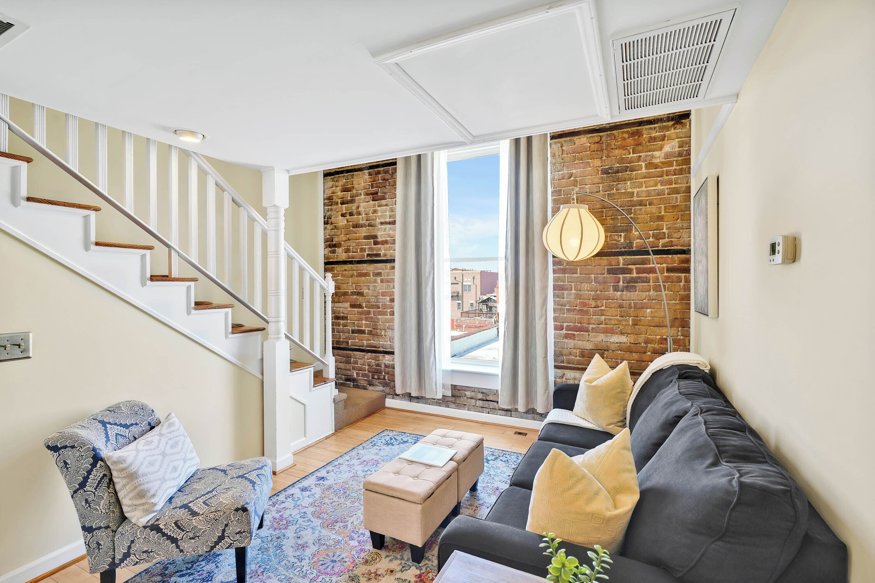 A living room with a staircase, a sofa, a chair, and a view through a large window.