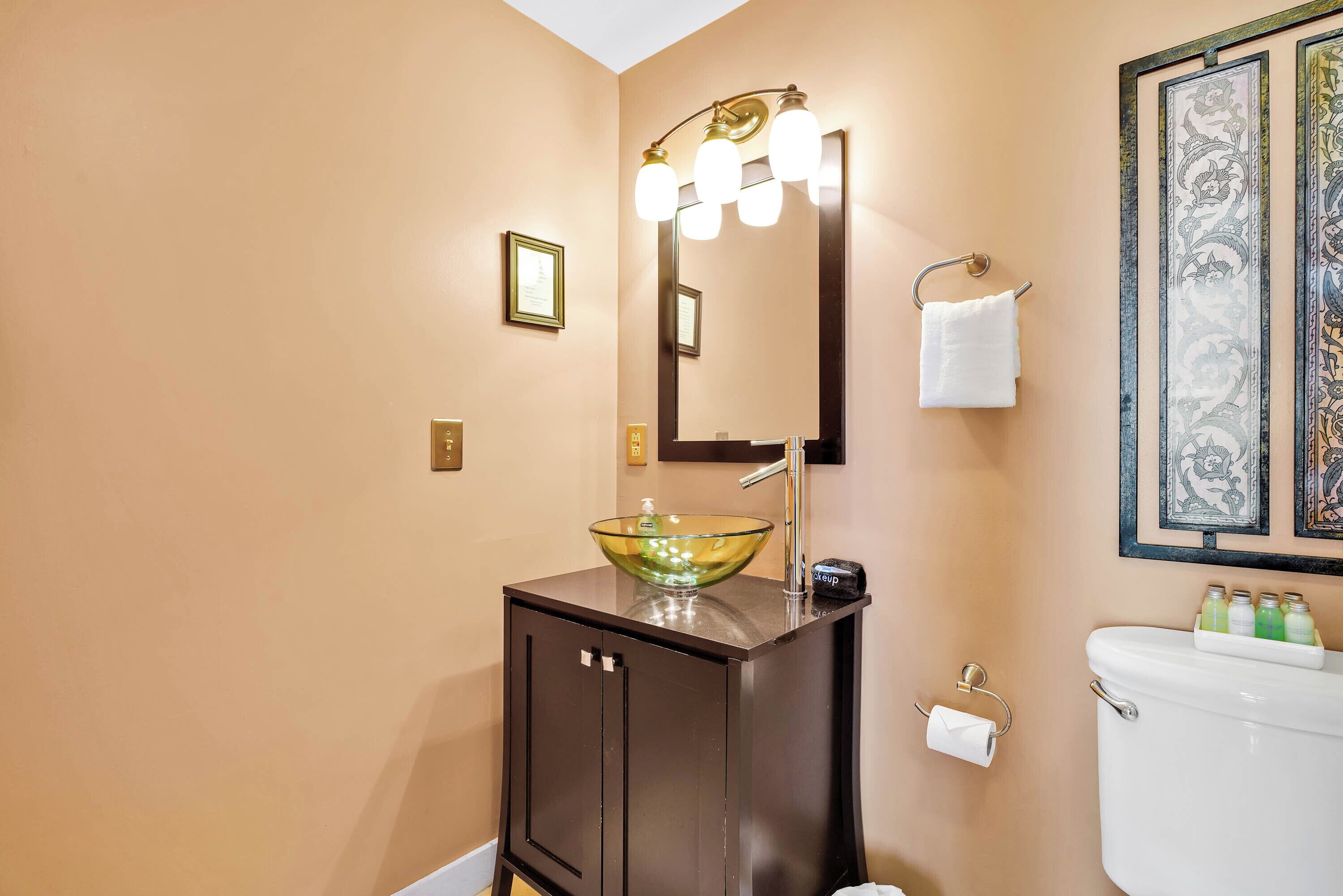 Une salle de bain avec un lavabo en verre, un meuble sombre, un miroir avec éclairage intégré et des œuvres d’art encadrées accrochées au mur.