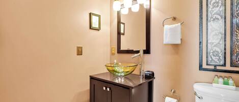 A bathroom with a glass bowl sink, a dark cabinet, a mirror with lights, and framed artwork on the wall.