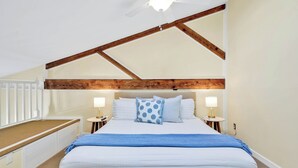 Ein Schlafzimmer mit geneigter Decke, Holzbalken, einem Bett mit weißer Bettwäsche, zwei Nachttischlampen und einer blauen Tagesdecke.