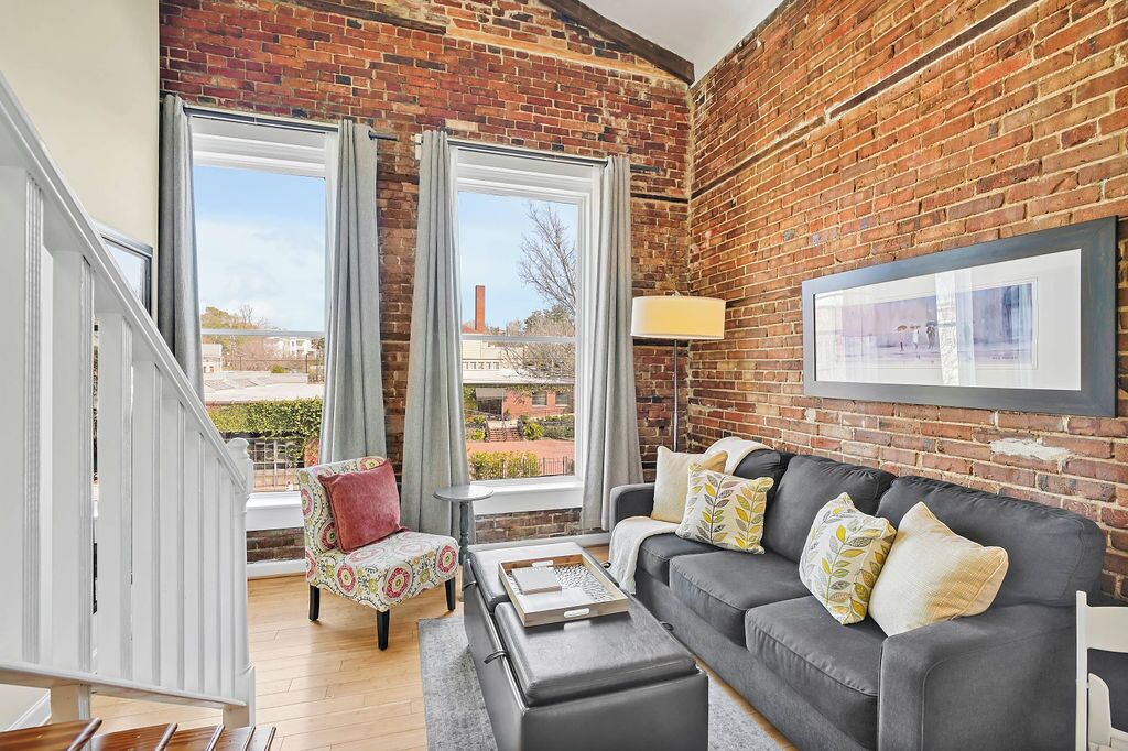 A living room with a brick wall, a grey sofa, a patterned armchair, and a large window with curtains.
