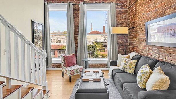 A spacious living area with a brick wall, a grey sofa with patterned cushions, a small table, and a chair with a floral cover. - Stemmerman Suite on Front! Perfect location in the Heart of Downtown! (wilmington)