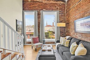 A spacious living area with a brick wall, a grey sofa with patterned cushions, a small table, and a chair with a floral cover. - Stemmerman Suite on Front! Perfect location in the Heart of Downtown! (wilmington)