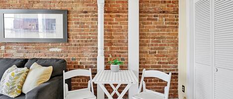 A room with a brick wall, a white table with two chairs, a grey sofa with cushions, and a framed picture on the wall.