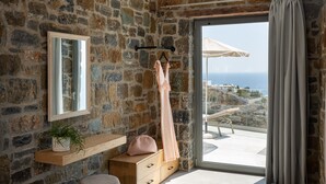 A stone-walled bathroom with a wooden vanity, a hanging robe, and a view of the sea through a large glass door.