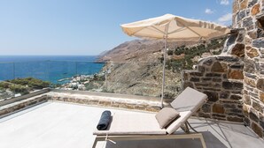 A balcony with a lounge chair and an umbrella overlooking a coastal landscape with a stone wall and a view of the sea.
