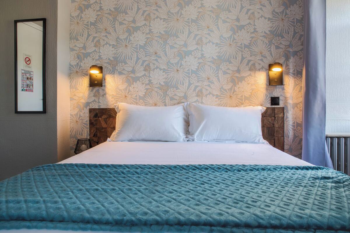A hotel room with a large bed, floral wallpaper, two wall-mounted lights, and a wooden headboard.